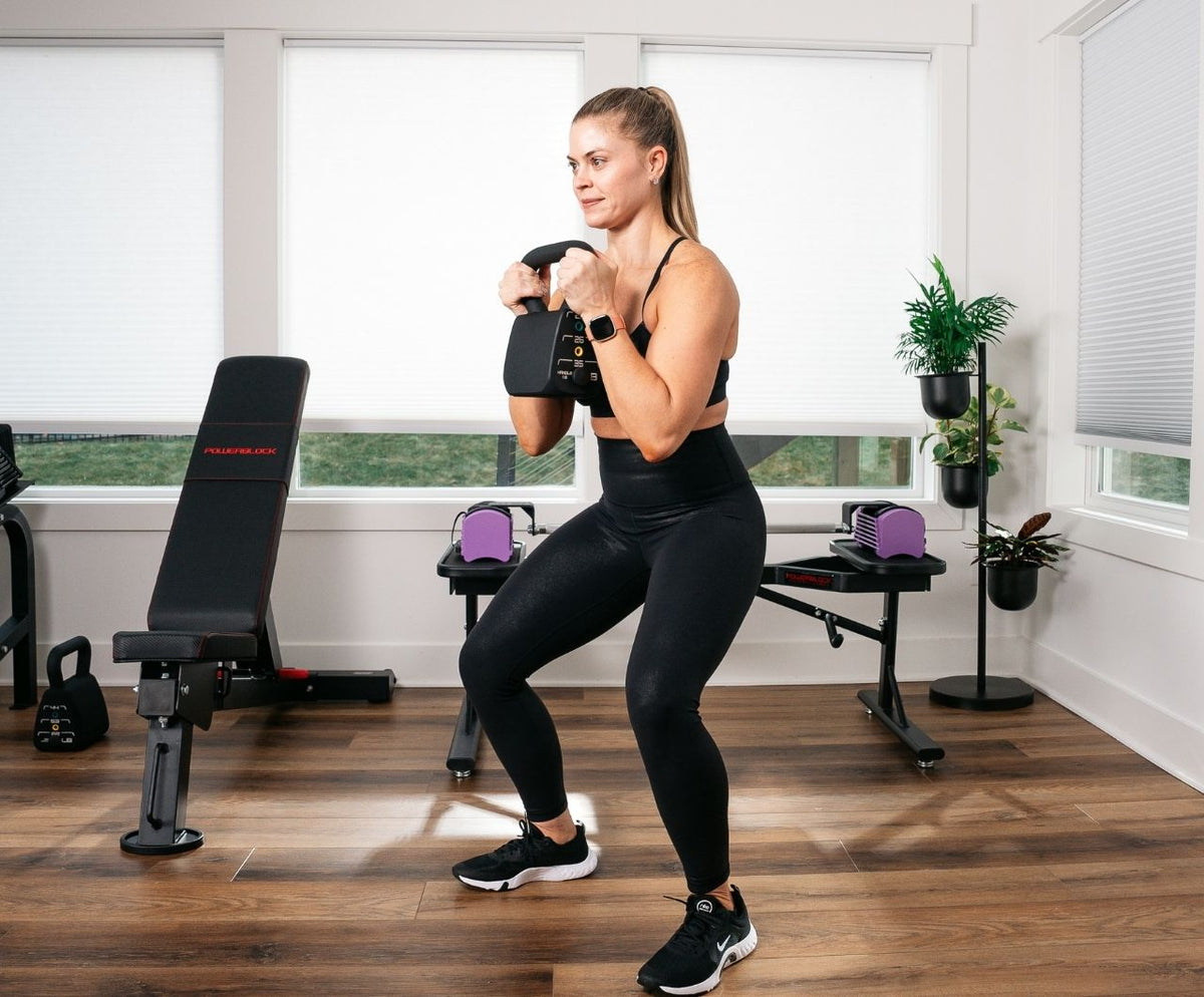 Woman performing goblet squats using the PowerBlock Pro Adjustable Kettlebell (18-35 lbs.) in front of the PowerBlock Ecosystem of Home Gym Equipment.