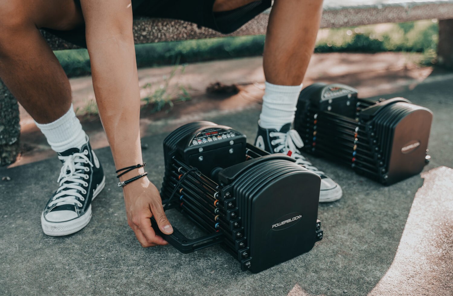 A person on an outdoor bench is bending down to make adjustments on one of two PowerBlock dumbbells.