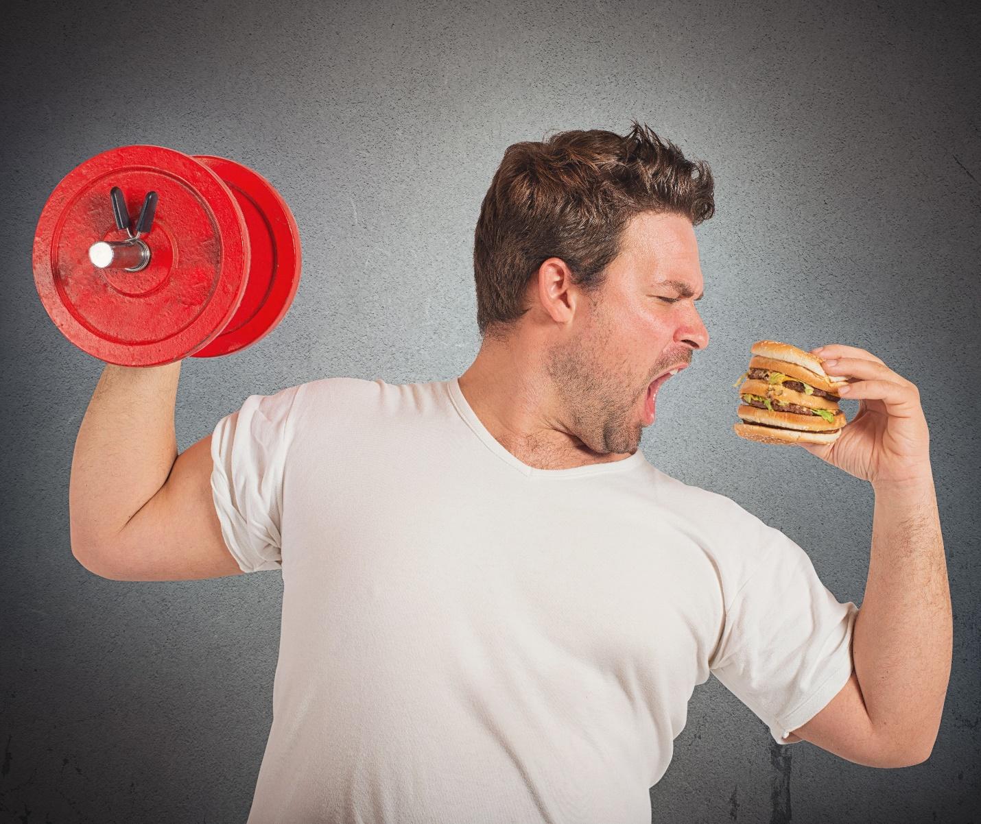 man holding a dumbbell and about to bite into a cheeseburger