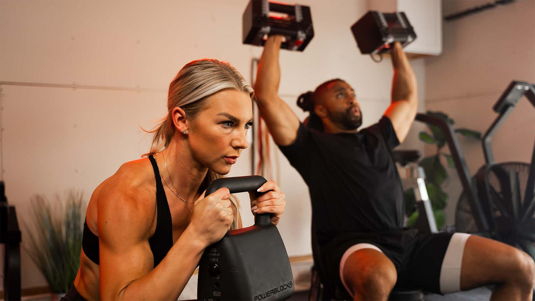 female athlete performs a goblet squat with a kettlebell while a male athlete performs a shoulder press with dumbbells in a home gym