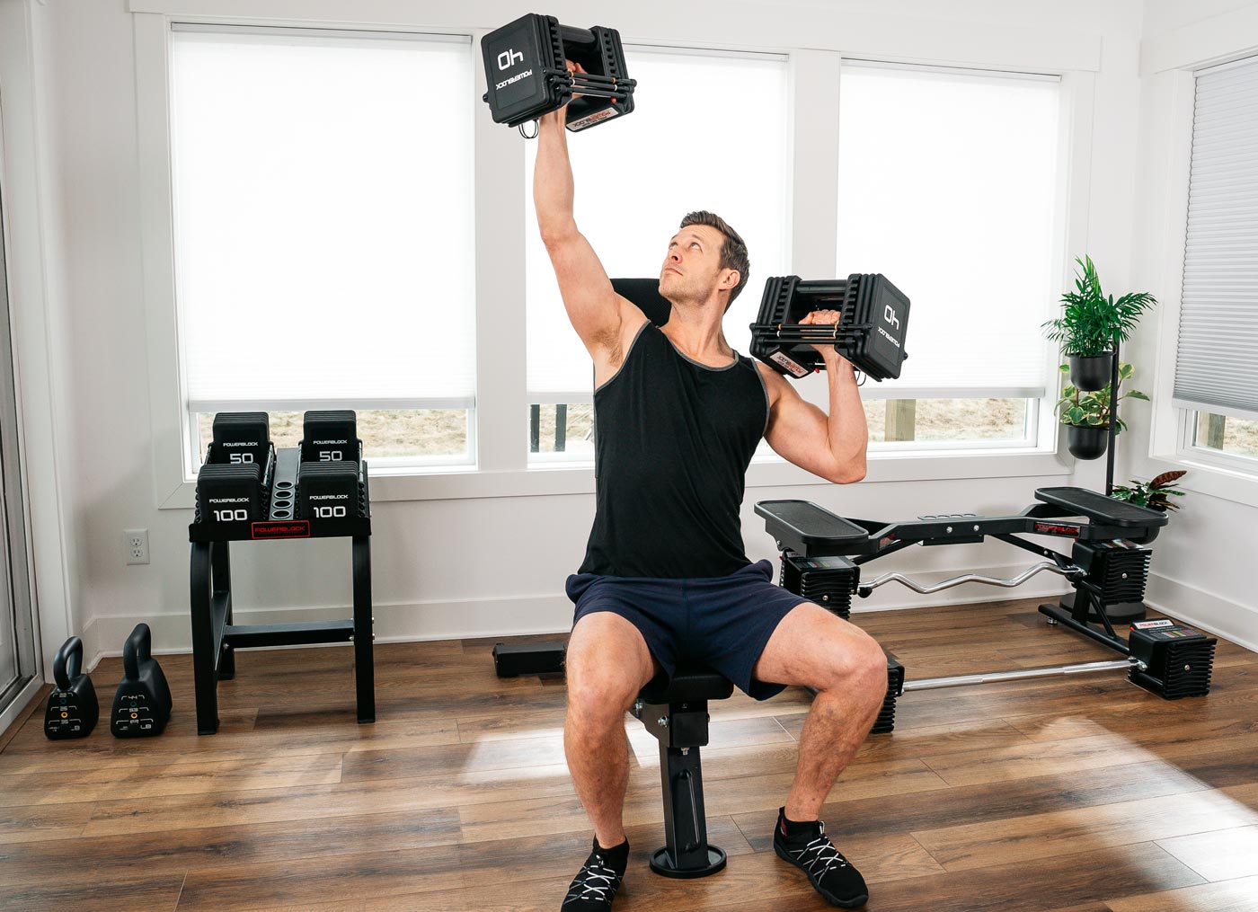 Man performing alternating shoulder presses using the Pro 100 EXP Adjustable Dumbbell and the PowerBench 2.0