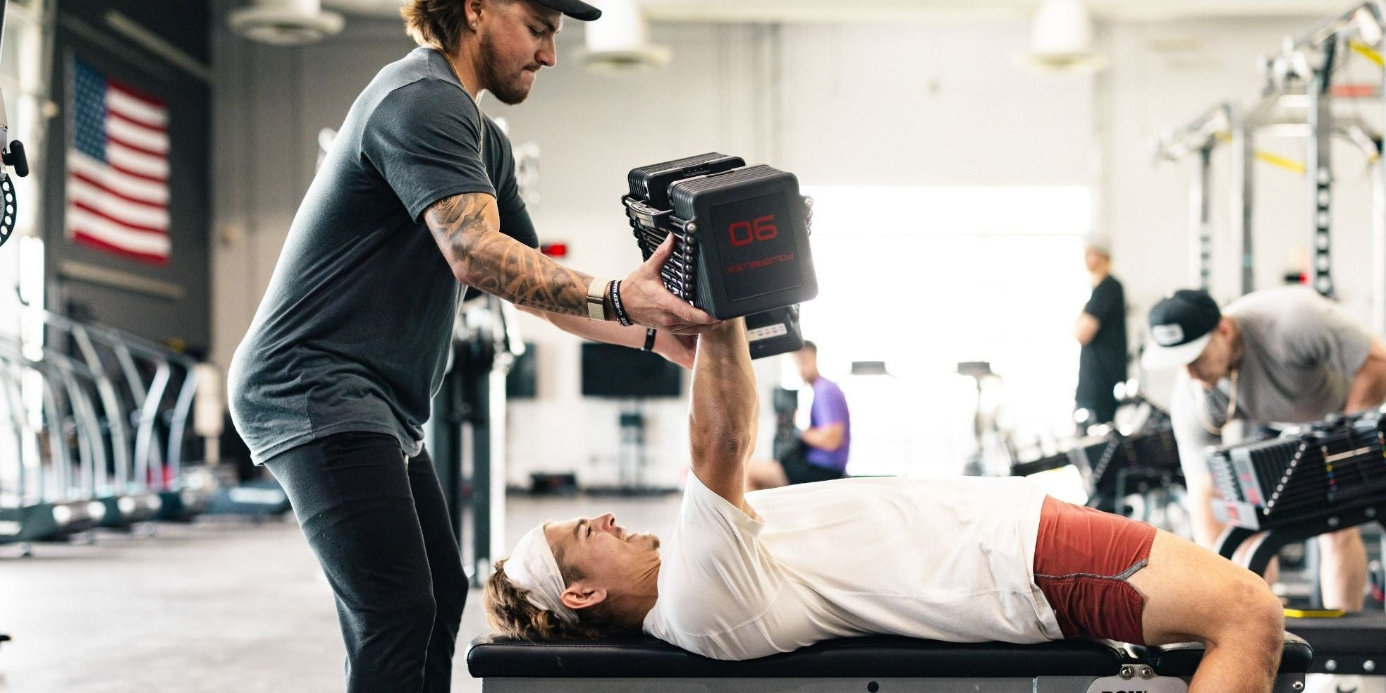 PowerBlock adjustable dumbbells being used in a professional training facility. One athlete performs a benchpress while the other spots.