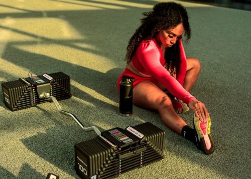 A woman stretching on the ground next to a PowerBlock Pro 100 EZ Curl Bar dumbbell attachment.