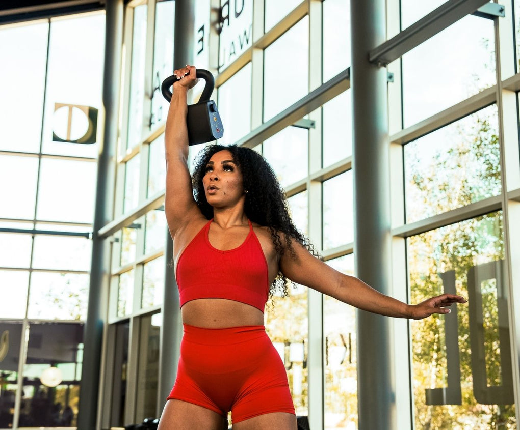 Woman in red athletic wear lifting a PowerBlock Adjustable kettlebell in front of large glass windows at the gym.