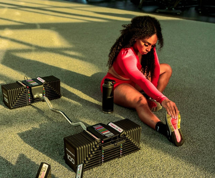 A woman stretching on the ground next to a PowerBlock Pro 100 EZ Curl Bar dumbbell attachment.