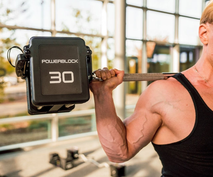 A close up of a man holding PowerBlock Pro 100 Straight Bar and Pro 100 EXP Adjustable Dumbbells on his shoulders while squatting.