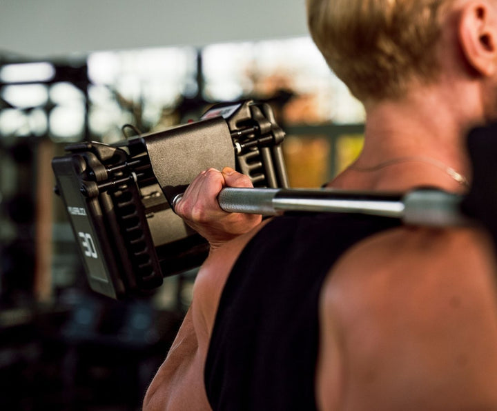 Man holding PowerBlock Pro 100 Straight Bar and Pro 100 EXP Adjustable Dumbbells on his shoulders.