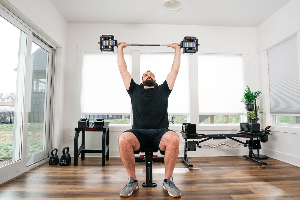male athlete performing a shoulder press with the PowerBlock adjustable barbell attachment with his Elite USA dumbbells showcasing how different PowerBlock equipment works together in a home gym.