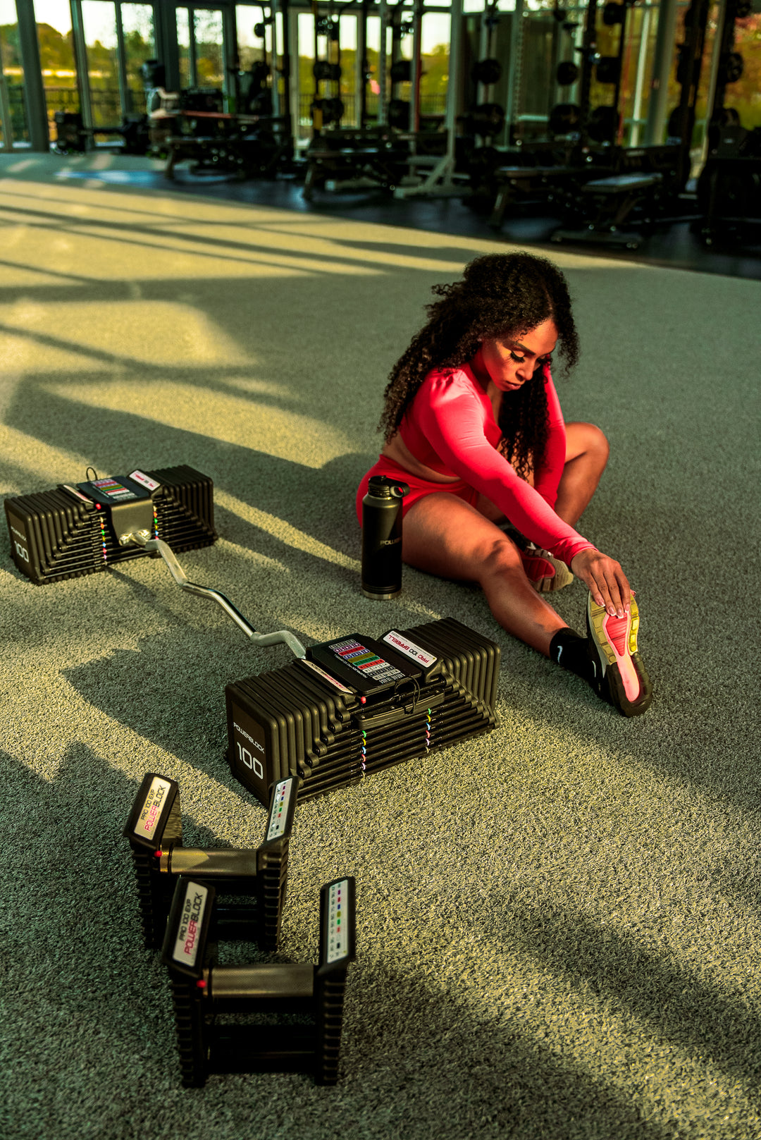 A woman stretching on the ground next to a PowerBlock Pro 100 EZ Curl Bar dumbbell attachment.
