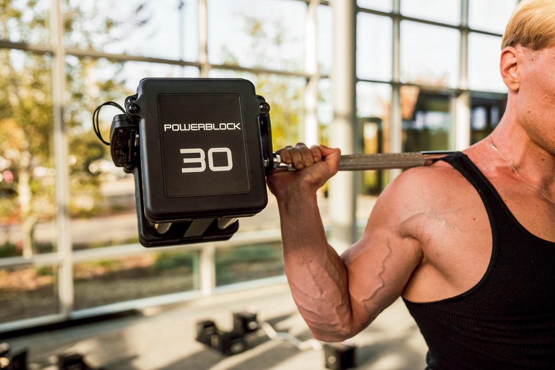 A close up of a man holding PowerBlock Pro 100 Straight Bar and Pro 100 EXP Adjustable Dumbbells on his shoulders while squatting.