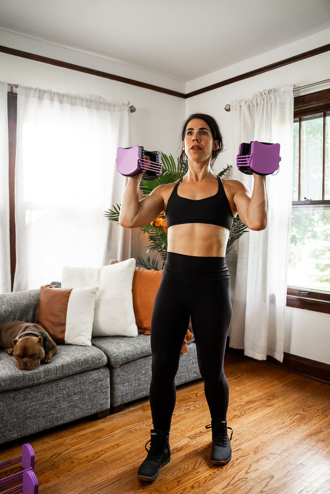A woman in her home using two PowerBlock lavender Sport 24 Adjustable Dumbbells for shoulder presses.