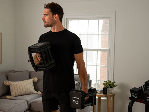 A man in his house doing bicep curls with a pair of PowerBlock Pro 100 EXP dumbbells set at 30 pounds infront of a PowerBlock home rack stand holding the remaining dumbbell weight plates