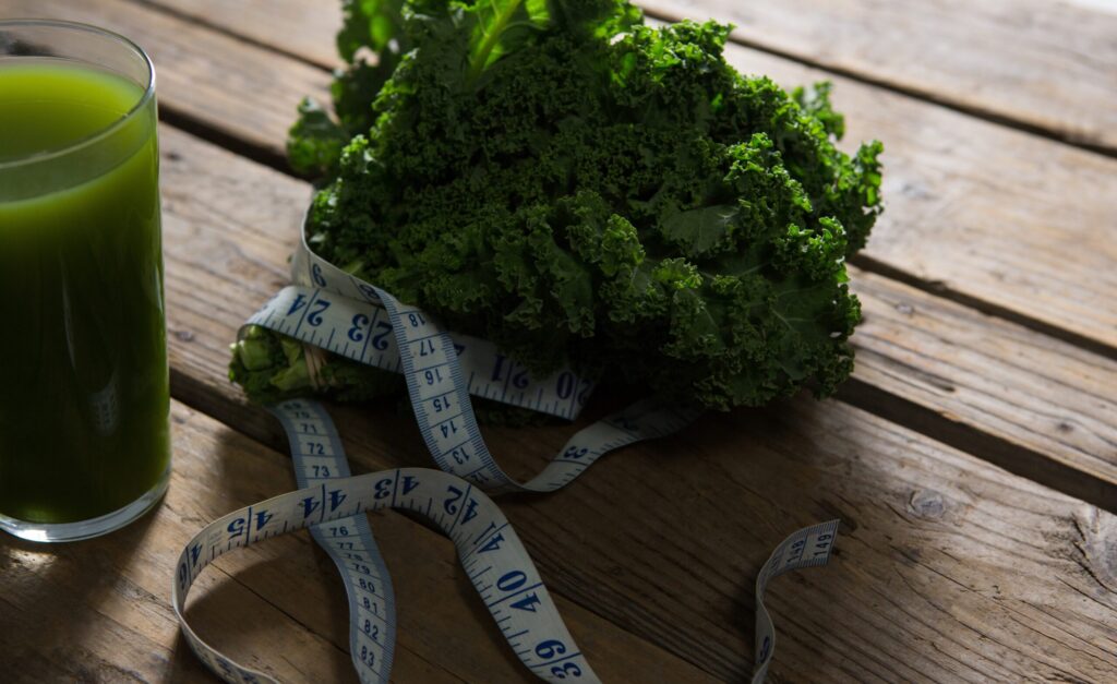 A large pile of broccoli on a tabletop, along with a tape measure and a glass of juice.