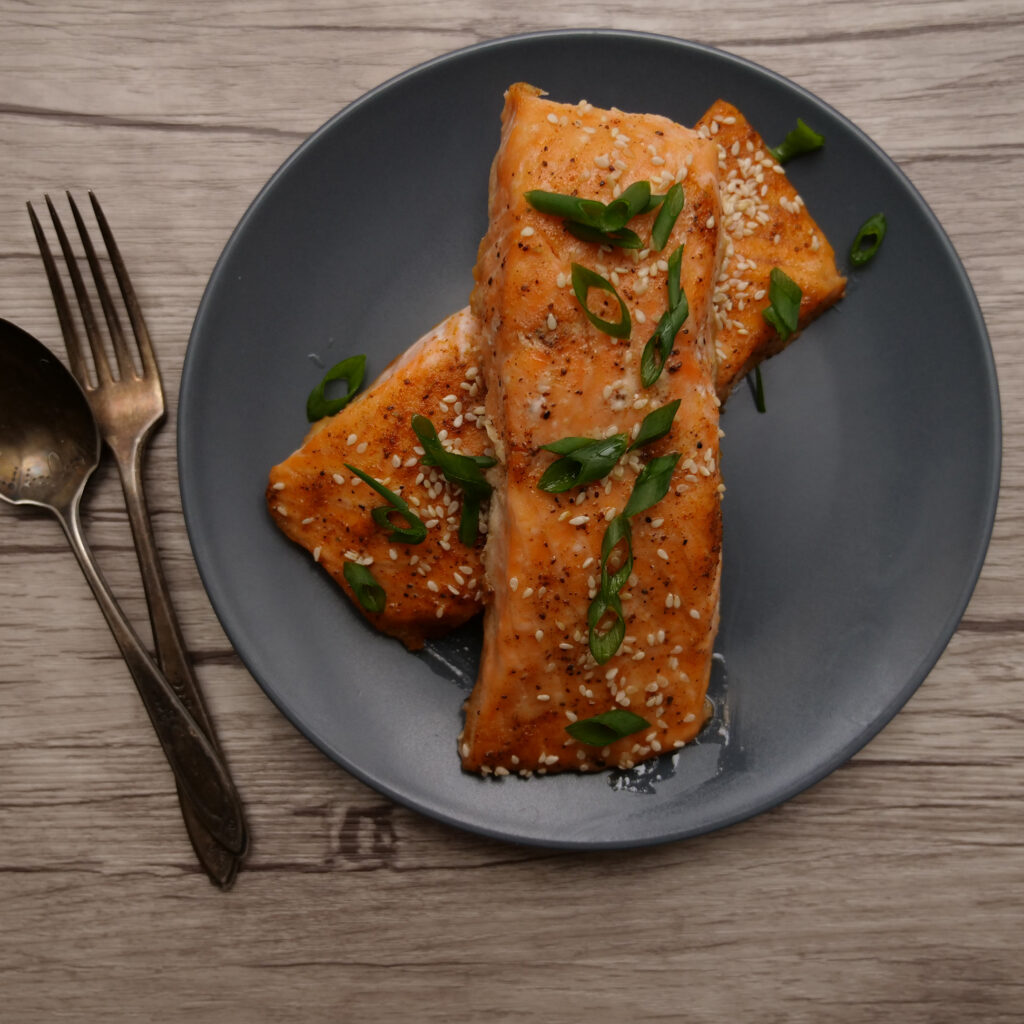 Two maple glazed salmon filets on a blue-gray plate, with a spoon and fork to the left.