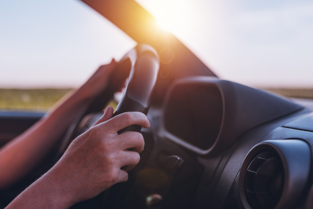 A pair of hands grip a steering wheel with an out-of-focus sunset or sunrise in the background.