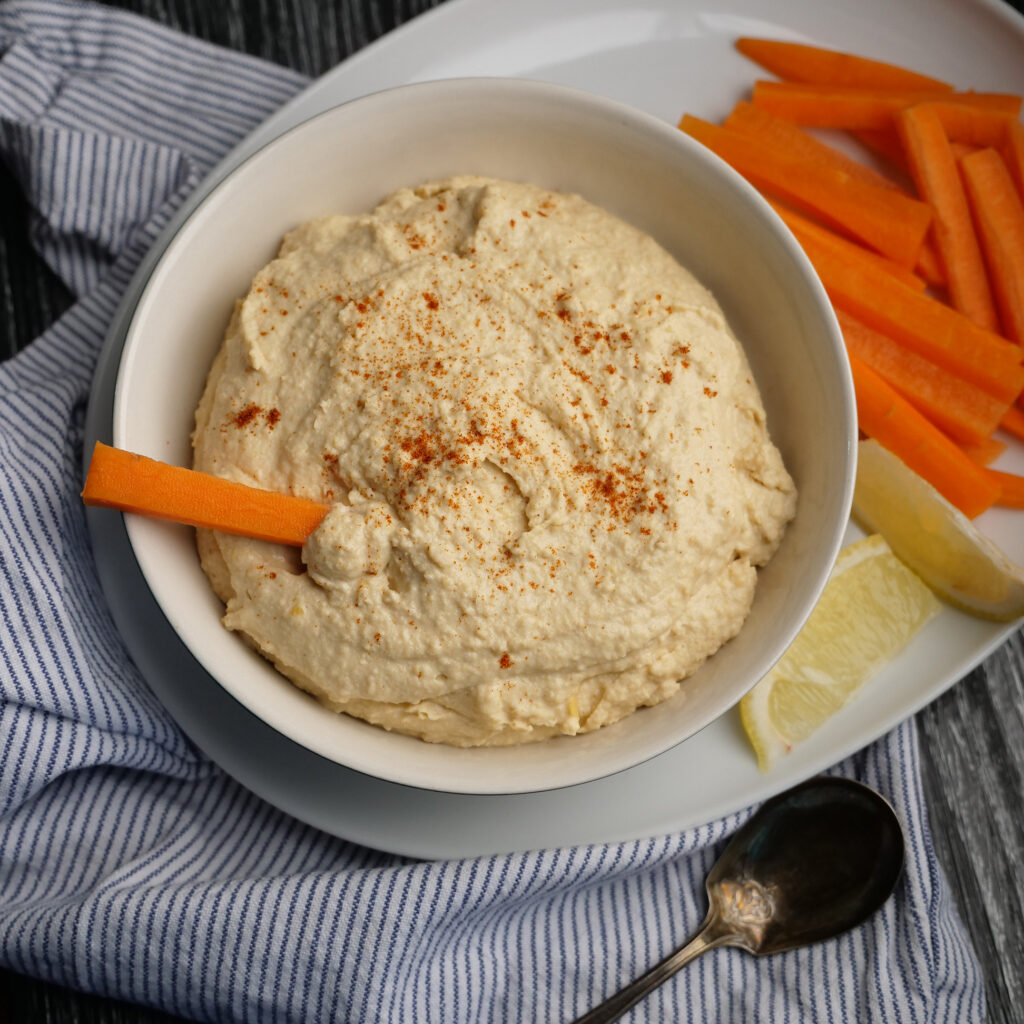 A bowl full of hummus, with carrots and lemon wedges adjacent.