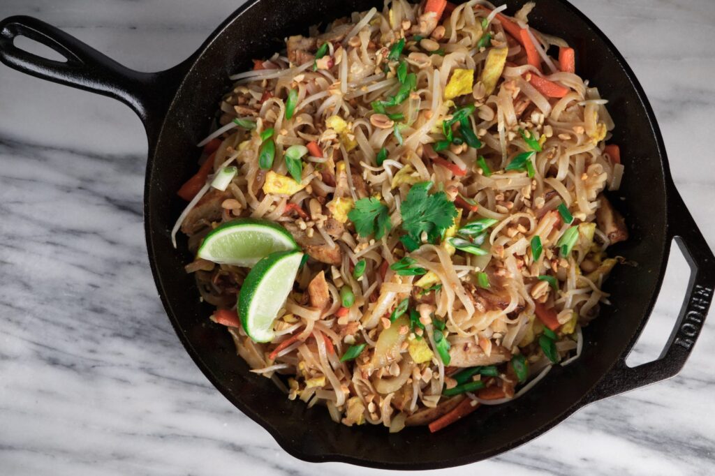 Cast iron skillet of Pad thai featuring noodles, vegetables, mint and lime, set on a marble surface.