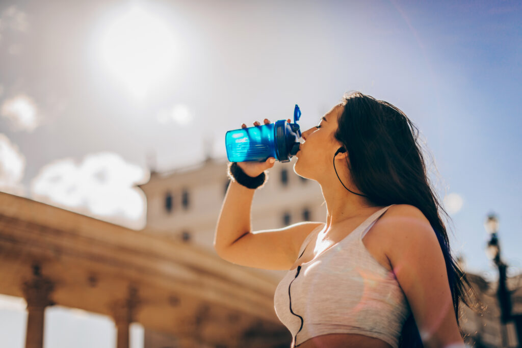 Upper half of a woman drinking from a water bottle with a building and sky background.