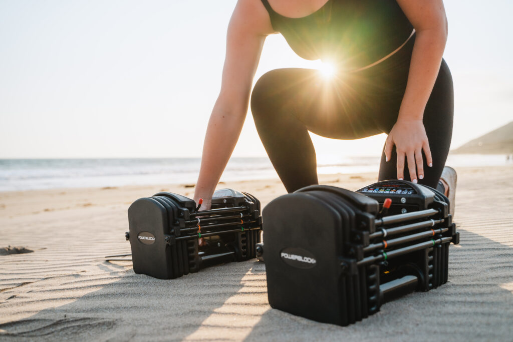 A woman kneeling on the beach with a hand grabbing one of a pair of adjustable PowerBlock dumbbells.