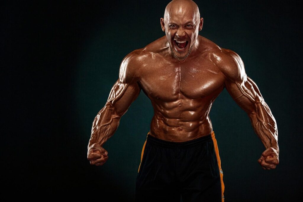A bald, shirtless man with large muscles yells into the camera.